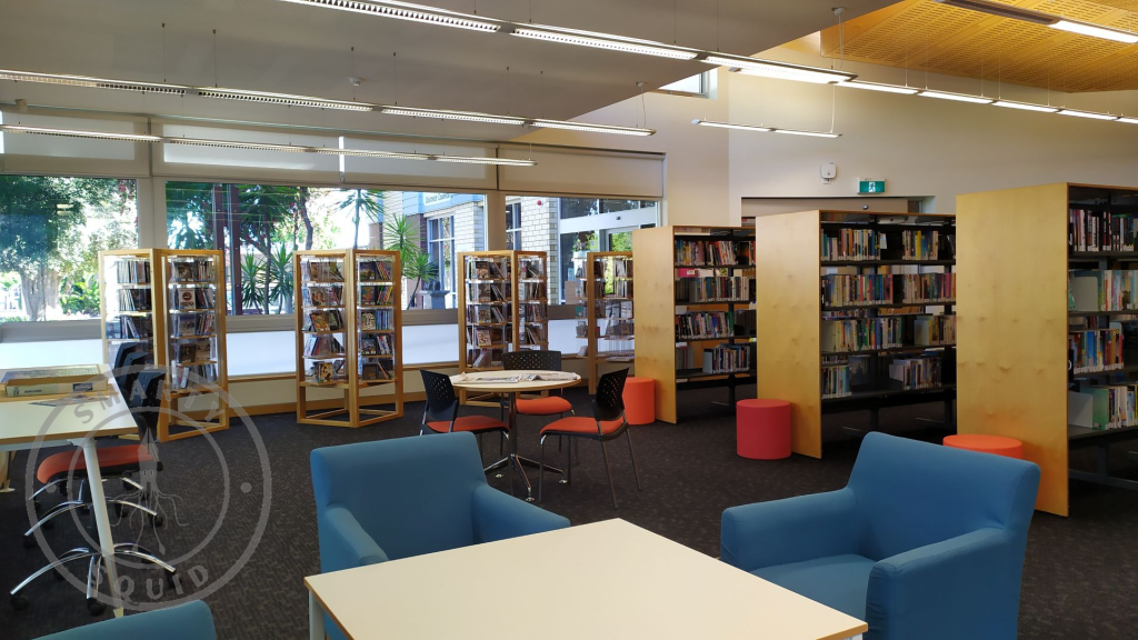 inside Bordertown Library looking at book shelves in the background and a table and two blue seats in the foreground

