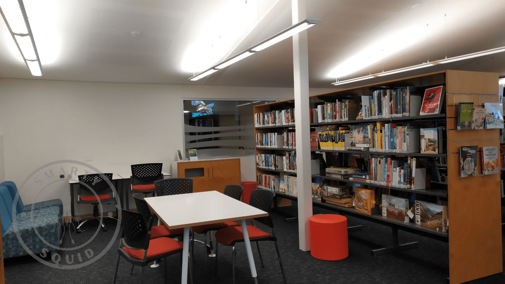 inside Bordertown Library looking at one row of shelves on the right and a table with red chairs in the middle
